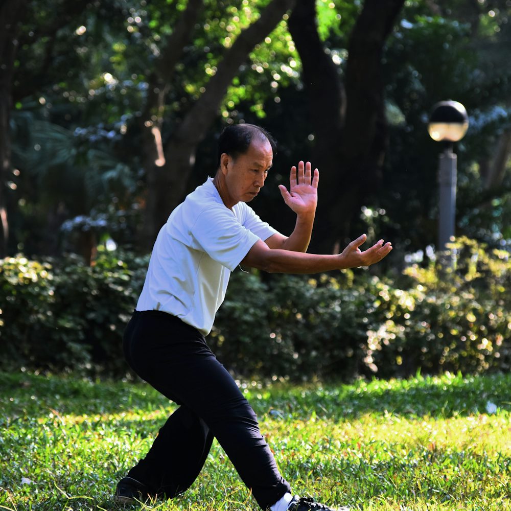 An older man in a white shirt practising tai chi among trees in a park
