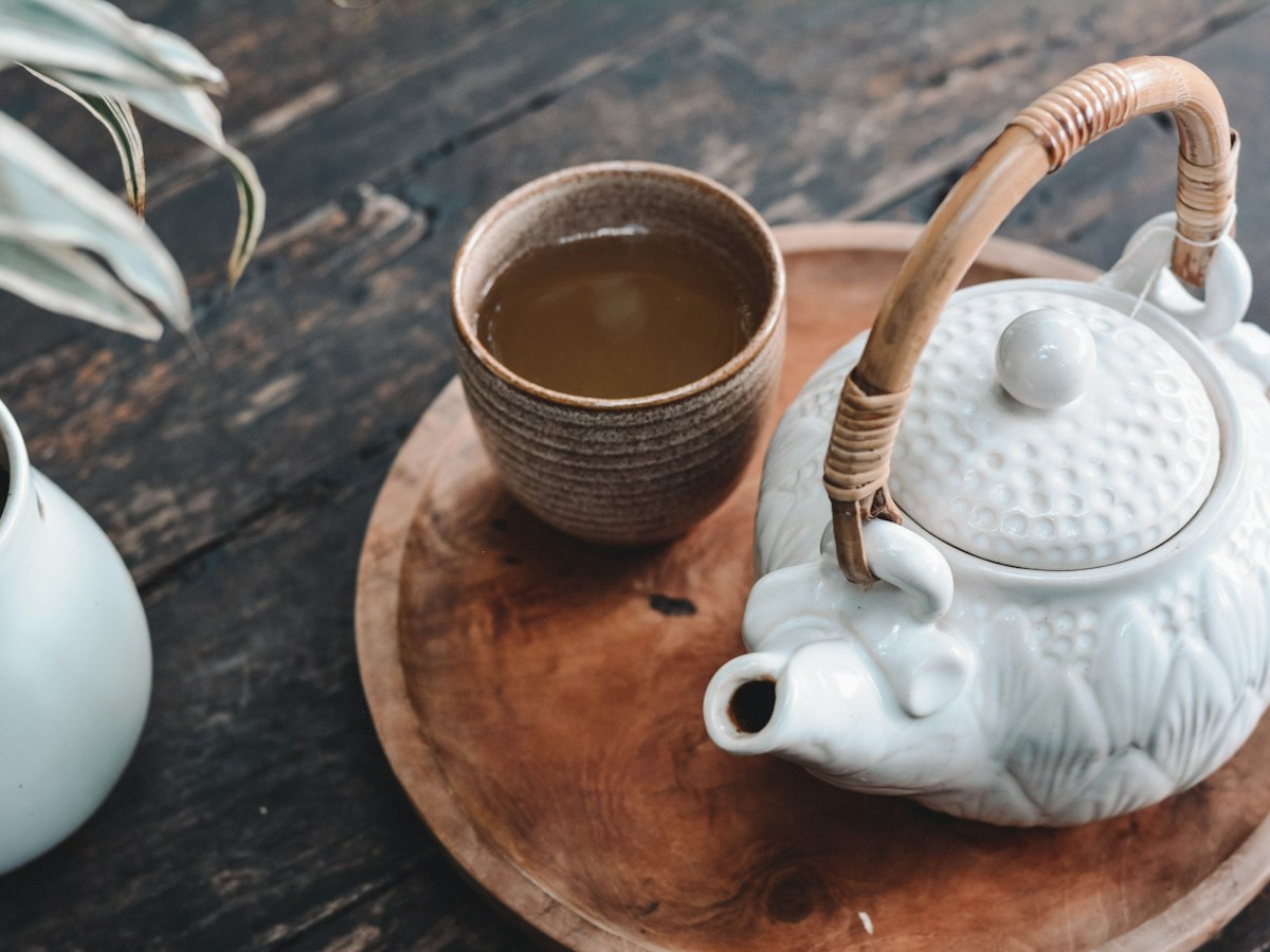 A white teapot with a cane handle and a cup of tea on a round wooden tray