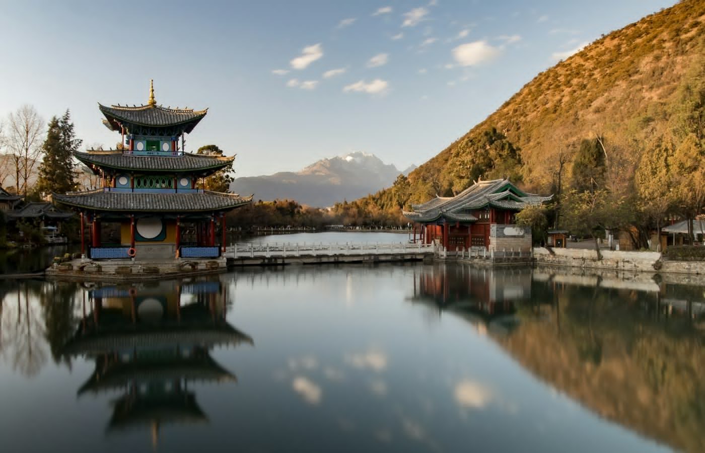 A traditional pavilion reflected in a calm lake, with a bridge and a snow-capped mountain behind